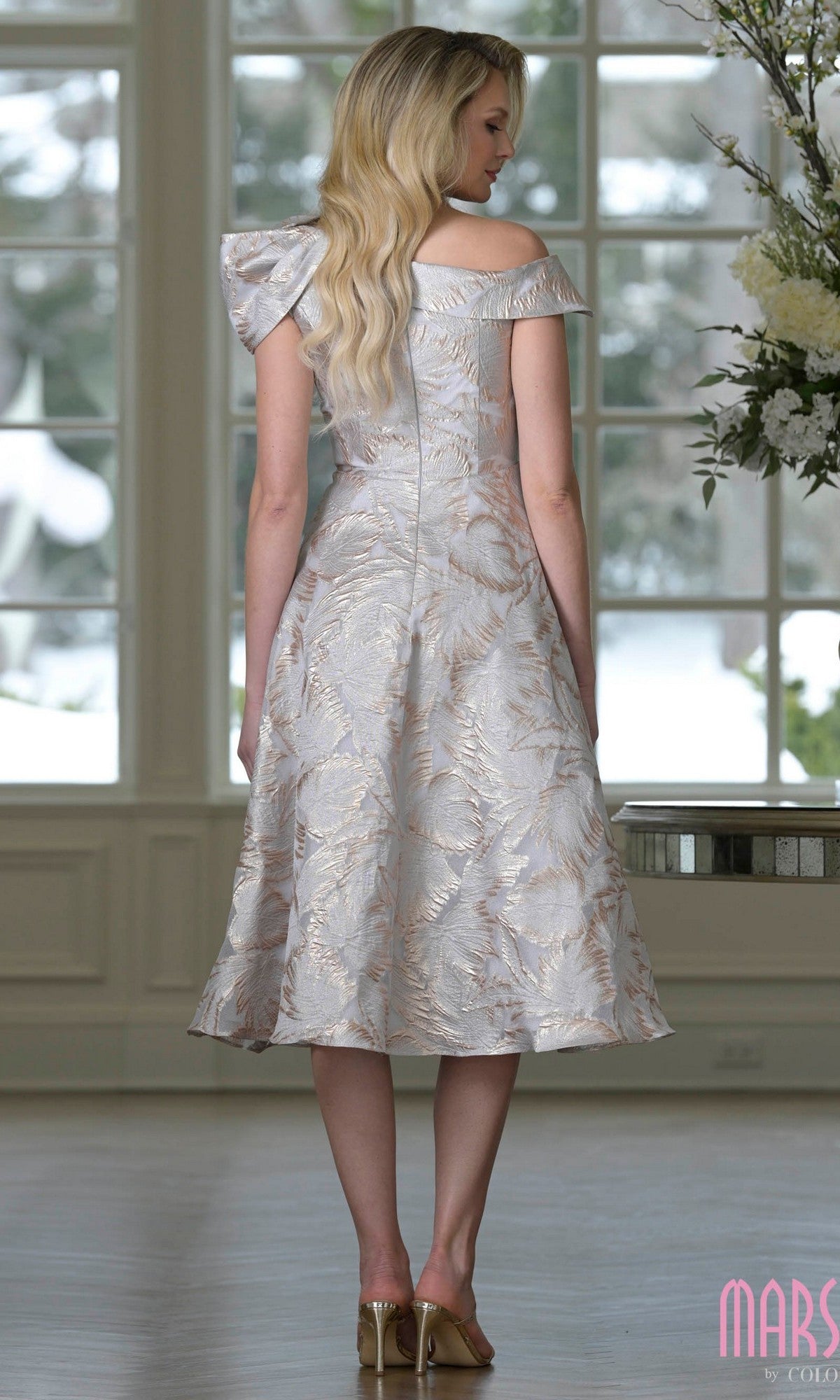 A woman with long blonde hair is wearing the Marsoni MV1413 knee-length semi-formal dress, featuring an off-the-shoulder design and subtle floral pattern, paired with high-heeled sandals and standing indoors facing away.